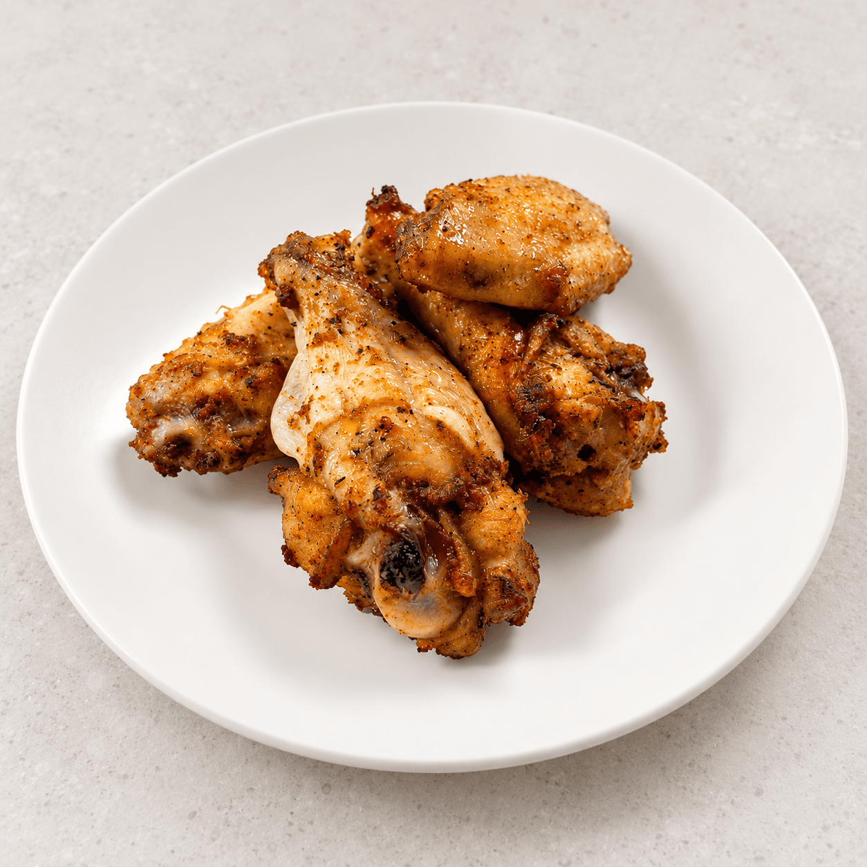 Four golden-brown, seasoned chicken wings arranged on a white plate on a light gray surface.